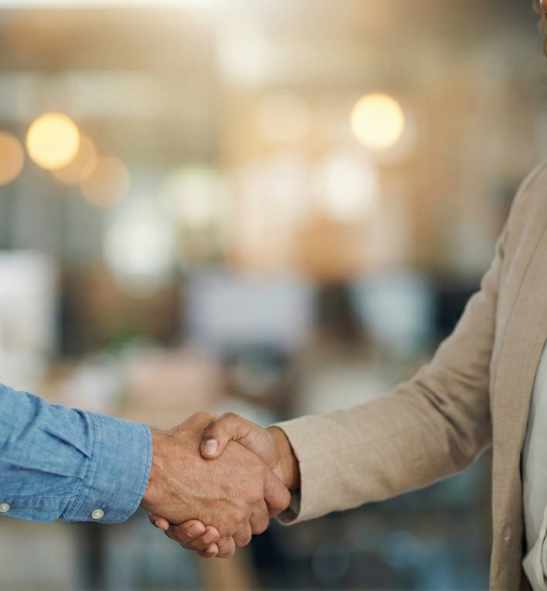 Symbolbild: Nahaufnahme eines Händedrucks zwischen zwei Personen in geschäftlicher Kleidung vor einem unscharfen Bürohintergrund. 